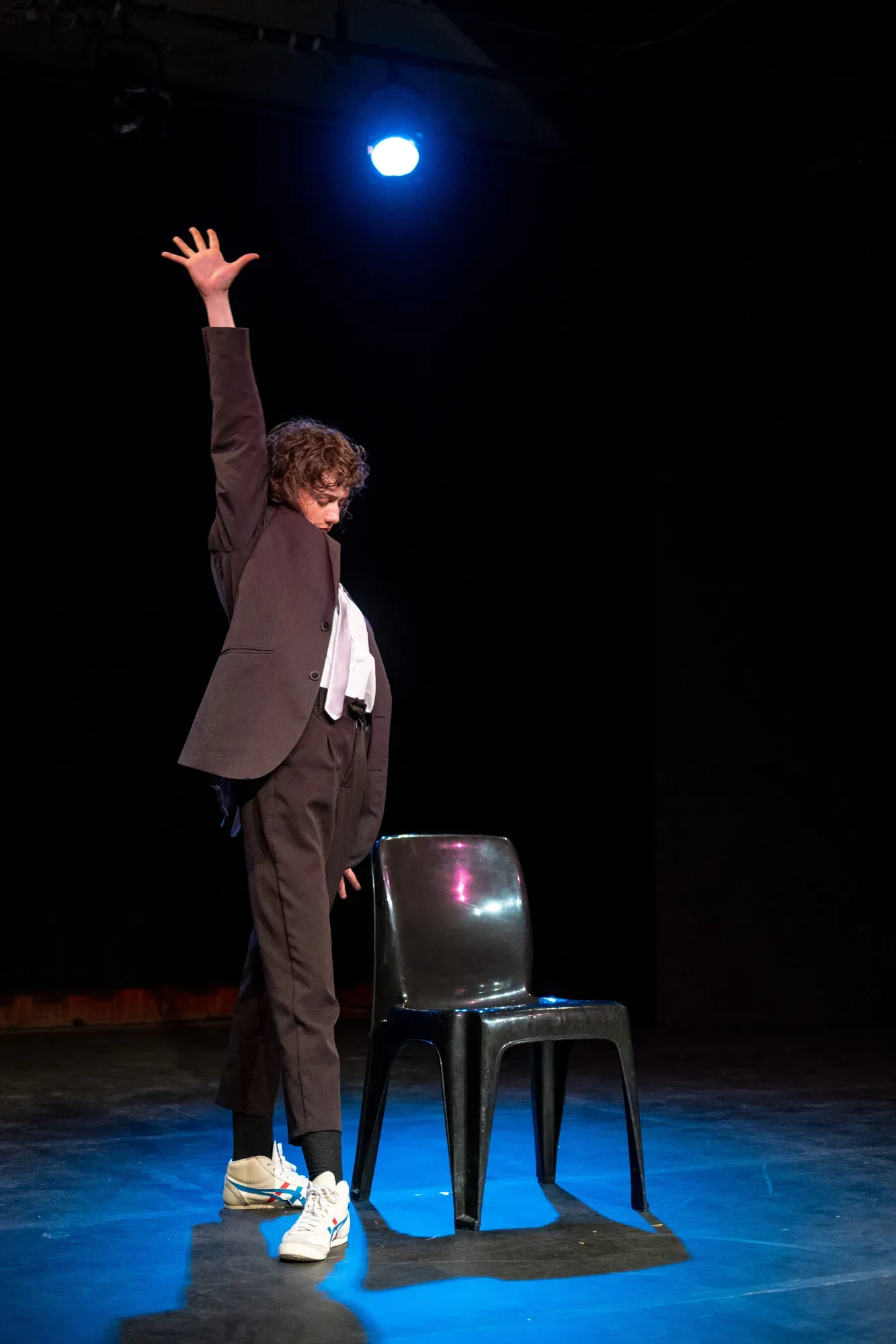 Person in a suit standing on a chair with one arm raised against a dark background